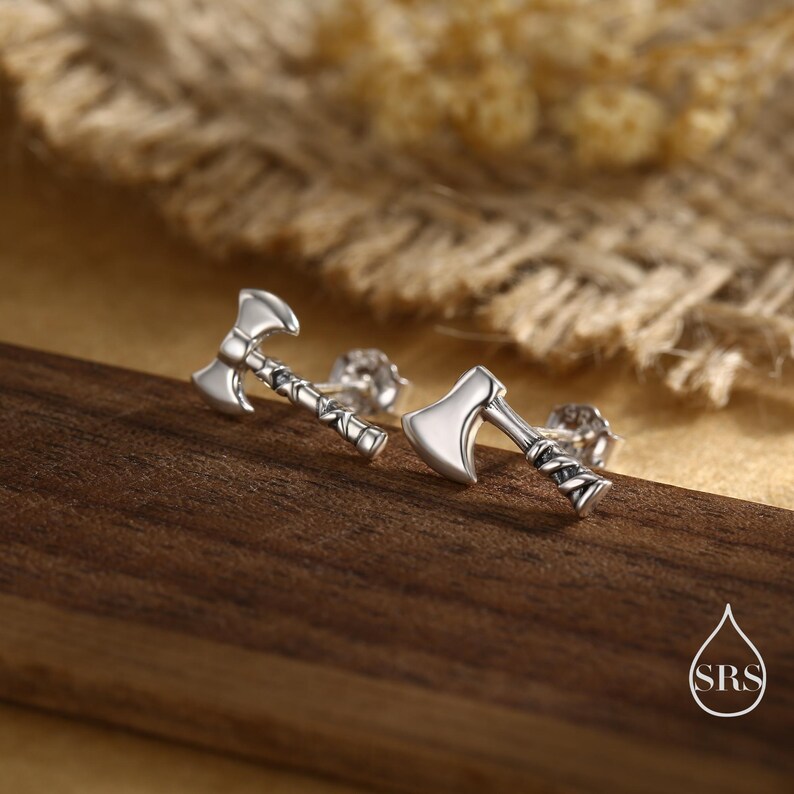 May include: Silver-colored axe-shaped stud earrings. The earrings are detailed with a textured handle and a polished axe head. The earrings are displayed on a wooden surface with a blurred background.