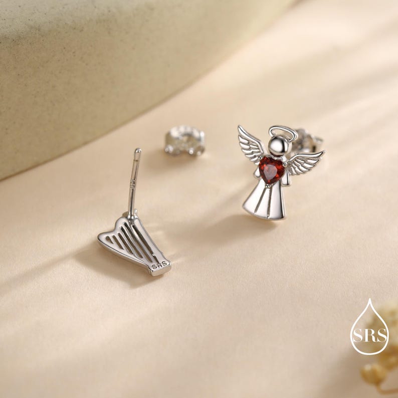May include: Silver angel earrings with a red heart-shaped gemstone. One earring is a complete angel design, the other is a harp-shaped earring back. The earrings are on a neutral background.