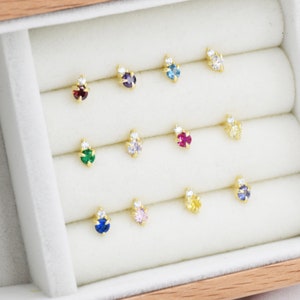 May include: A display of 12 small, gold-toned earrings with different colored gemstones. Each earring features a small, clear gemstone next to a larger, colored gemstone. The earrings are arranged in rows in a white velvet-lined box.