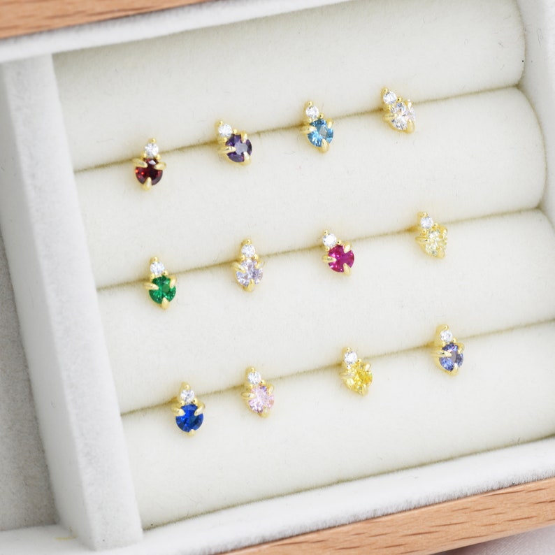 May include: A display of 12 small, gold-toned earrings with different colored gemstones. Each earring features a small, clear gemstone next to a larger, colored gemstone. The earrings are arranged in rows in a white velvet-lined box.