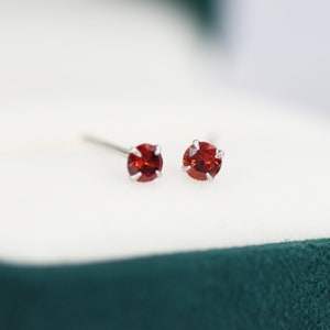 May include: A pair of small, silver stud earrings with red gemstones. The earrings are in a white box with a green velvet lining.