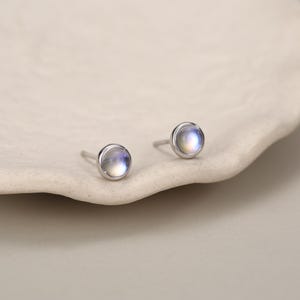 Puede incluir: Un par de pendientes de botón con piedras de luna redondas talladas en cabujón, engastadas en metal plateado pulido. Cada pendiente tiene una superficie lisa y abovedada que muestra un sutil brillo azul iridiscente, con cierre de poste.