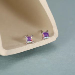 May include: A pair of stud earrings featuring square-cut iridescent stones that display shifting purple, blue, and pink hues. Each stone is held in a four-prong silver-toned metal setting, creating a reflective and multifaceted appearance.