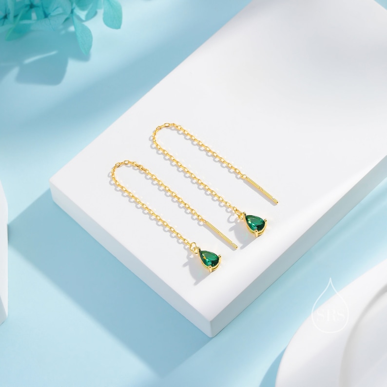 May include: A pair of gold-colored threader earrings. Each earring features a delicate chain, a small gold bar, and a teardrop-shaped, emerald-green gemstone. The earrings are displayed on a white surface with a light blue background.