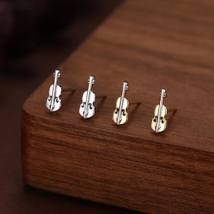 Puede incluir: Cuatro pares de pendientes con forma de violín. Dos pares son plateados y dos dorados. Los pendientes se exhiben sobre una superficie de madera oscura, mostrando su delicado diseño y tema musical.
