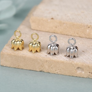 May include: Four small, flower-shaped jewellery findings. Two are gold-toned, and two are silver-toned. Each has a loop at the top for attaching to a chain or other jewellery component. The findings are on a light brown surface.