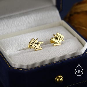May include: A pair of gold-colored stud earrings shaped like plague doctor masks. The earrings are displayed in a blue velvet jewelry box with a white interior. The earrings have a hat, beak, and eye details.