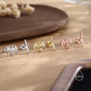 Peut inclure: Une collection de boucles d'oreilles &agrave; clous en argent, or et or rose. Les boucles d'oreilles pr&eacute;sentent des motifs de dragon et d'&eacute;p&eacute;e. Les boucles d'oreilles sont pr&eacute;sent&eacute;es sur une surface marron clair avec un arri&egrave;re-plan flou.
