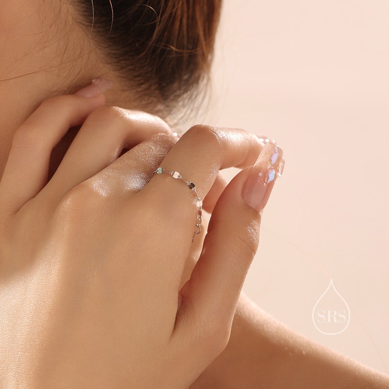 May include: A silver ring with a delicate chain and small round charms. The ring is worn on the index finger of a hand.