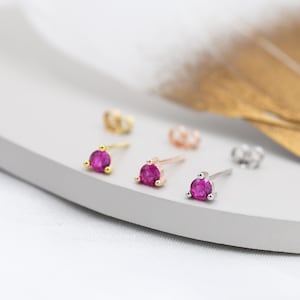 May include: Close-up of several stud earrings with bright pink gemstones. The earrings are set in gold, rose gold, and silver. The earring backs are also visible. The earrings are displayed on a white and grey surface.