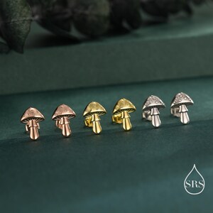 May include: A collection of mushroom-shaped stud earrings in rose gold, gold, and silver tones. The earrings are arranged in a row, showcasing their detailed design. The background is a dark green, creating a contrast that highlights the jewelry.