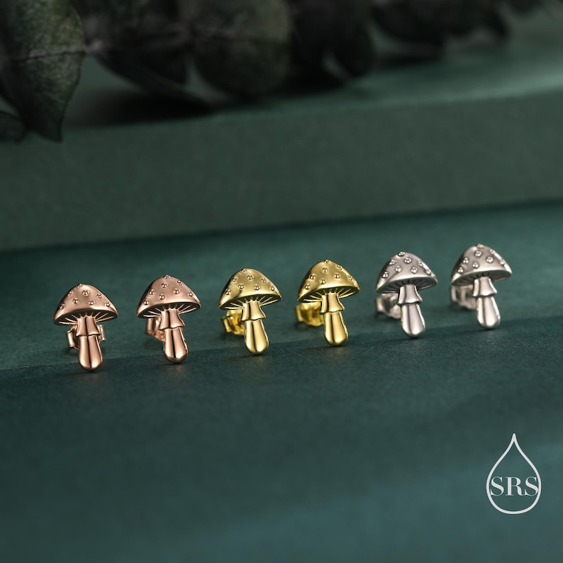 May include: A collection of mushroom-shaped stud earrings in rose gold, gold, and silver tones. The earrings are arranged in a row, showcasing their detailed design. The background is a dark green, creating a contrast that highlights the jewelry.