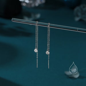 Puede incluir: Un par de pendientes de cadena plateada con un disco martillado y una barra recta. Los pendientes se exhiben en una varilla transparente sobre un fondo verde azulado. Diseño minimalista y elegante.