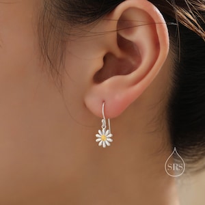May include: Silver dangle earrings with a white daisy flower charm and a yellow center. The earrings are shown on a person's ear.