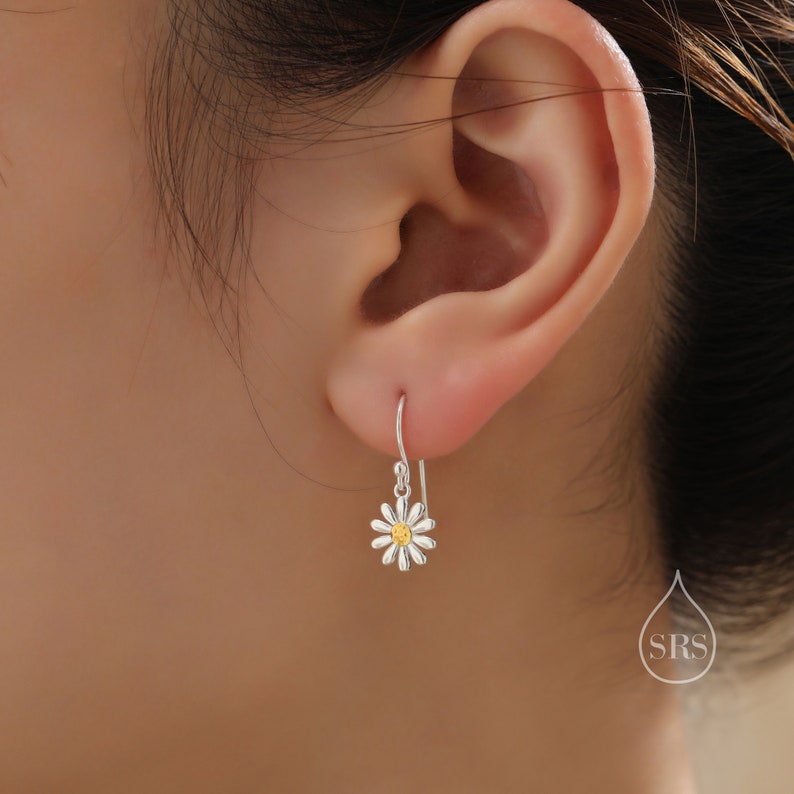 May include: Silver dangle earrings with a white daisy flower charm and a yellow center. The earrings are shown on a person's ear.