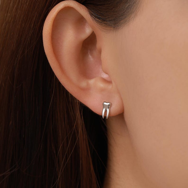 May include: A small, polished silver-toned stud earring shaped like a human tooth with two distinct roots. The earring is secured in the earlobe, featuring a smooth, reflective metallic surface that catches the light with a simple, clean design.