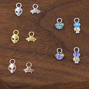 May include: A collection of small, metallic charms in various designs. The charms include alien heads, UFOs, planets with stars, and astronaut figures. The charms are in silver, gold, and rose gold tones, with some featuring iridescent blue and purple accents.