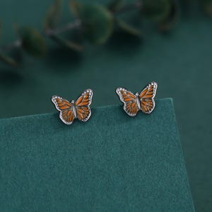 以下が含まれることがあります： 蝶のスタッドピアス。各イヤリングは、オレンジ色のエナメルウィングと白い点線ボーダーを備えたシルバーカラーの金属の輪郭が特徴です。イヤリングは、ターコイズグリーンの表面に表示されています。