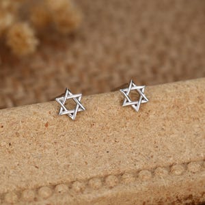 May include: A pair of silver stud earrings, each in the shape of a Star of David. The earrings are presented on a light brown surface. The earrings are small and delicate, with a simple, geometric design.