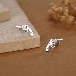 以下が含まれることがあります： 銀色のリボルバー型スタッドピアス。ピアスは銃身、シリンダー、ハンドルで詳細に表現されています。ピアスは、茶色の背景に対して、明るい茶色の表面に表示されています。