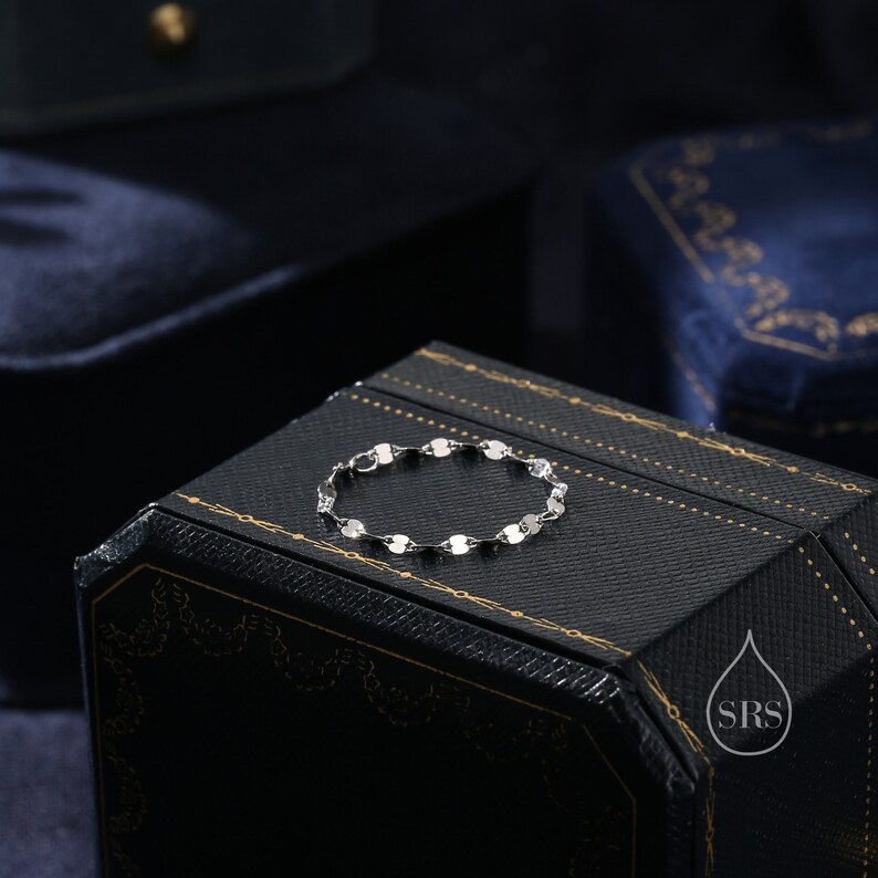 May include: A silver bracelet with a chain made of small, round links. The bracelet is lying on a black box with gold accents and the text "SRS" in a water drop logo.