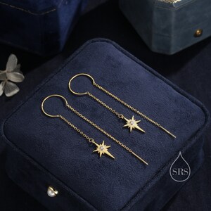 May include: A pair of gold dangle earrings with a star charm and a small clear stone in the center. The earrings are on a blue velvet background.