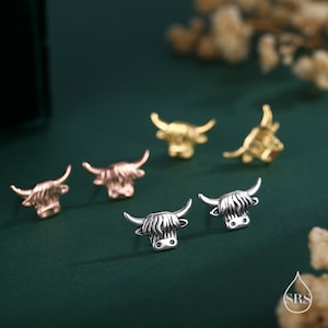 May include: A collection of Highland cow-shaped stud earrings in silver, rose gold, and gold tones. The earrings are displayed on a dark green surface, showcasing their detailed design and textured finish. The earrings are small and delicate.