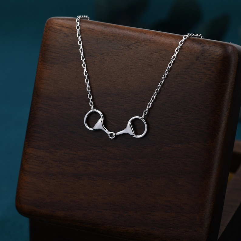 May include: A silver necklace with a simple, elegant design featuring two connected horse bit charms. The necklace is displayed on a dark brown wooden surface.