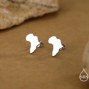 May include: A pair of silver stud earrings in the shape of the African continent. The earrings are presented on a brown surface, with a blurred background. The earrings are small and shiny.