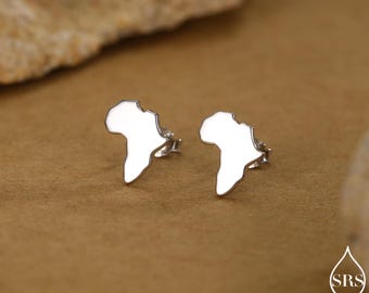 May include: A pair of silver-colored stud earrings shaped like the African continent. The earrings are displayed on a brown surface, with a blurred background. The earrings are small and shiny.