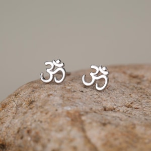 May include: A pair of silver-toned stud earrings, each featuring the Om symbol. The earrings have a polished, metallic finish and are designed as small, delicate shapes that represent the sacred sound and spiritual icon in a simple, clean form.