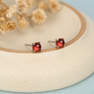 Puede incluir: Un par de pendientes de botón con pequeñas piedras preciosas rojas, redondas y translúcidas, engastadas en garras de metal plateado. Los pendientes están colocados sobre una superficie circular de color beige claro con textura.
