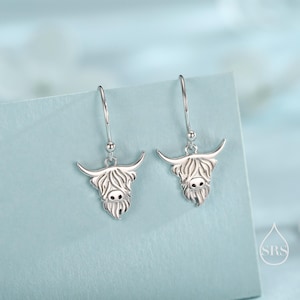 Puede incluir: Pendientes colgantes plateados con diseños de vaca de las Tierras Altas. Cada pendiente muestra una cabeza de vaca detallada con cuernos, una cara esponjosa y pequeños ojos negros. Los pendientes cuelgan de ganchos simples.