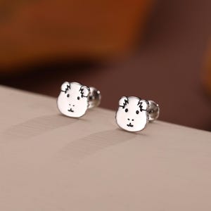 May include: A pair of silver-colored stud earrings. Each earring features a white, bear-shaped design with black detailing for the eyes, nose, and mouth. The earrings are set against a neutral background.