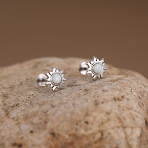 May include: A pair of silver stud earrings. Each earring features a sunburst design with a central, round, white opal stone. The earrings are displayed on a textured, light brown surface.