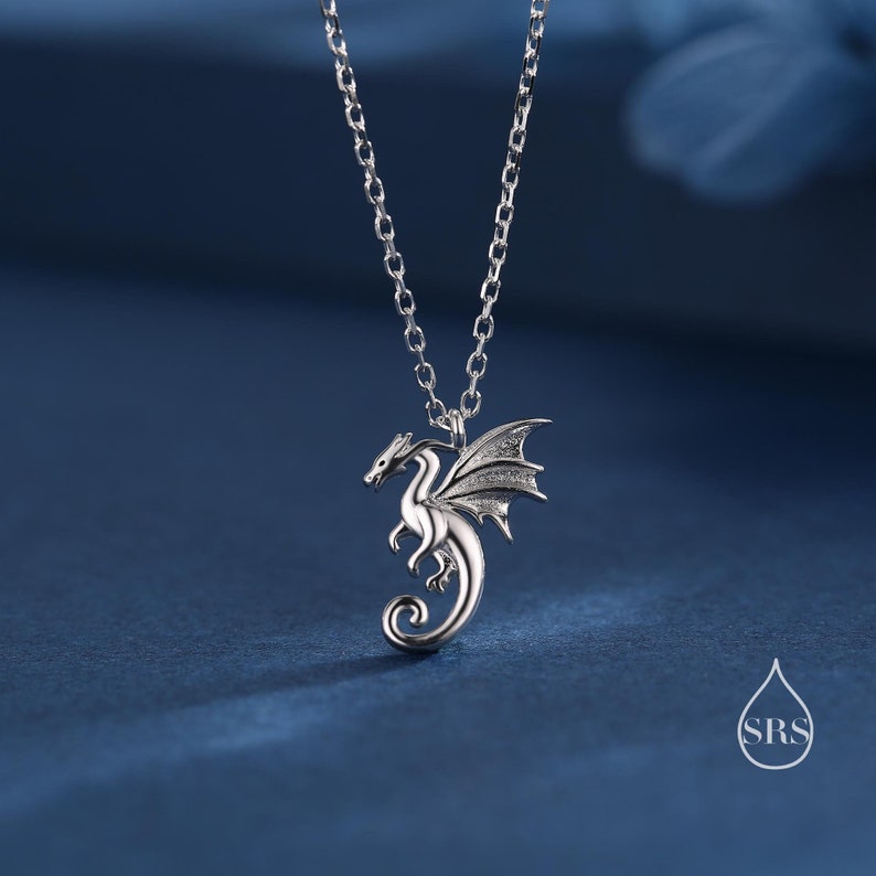 May include: A silver-toned metal necklace featuring a pendant shaped like a dragon with spread wings and a curled tail. The pendant hangs from a delicate silver-toned chain. A small white logo with the letters SRS inside a drop shape is visible.