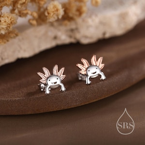 May include: Silver axolotl stud earrings with rose gold detailing. The earrings depict a smiling axolotl, with detailed gills and a cheerful expression. A unique and charming accessory for everyday wear.