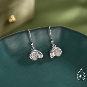 May include: A pair of silver-colored drop earrings. Each earring features a detailed, openwork design resembling a seashell. The earrings hang from a simple hook. The earrings are displayed on a teal-colored ceramic plate.