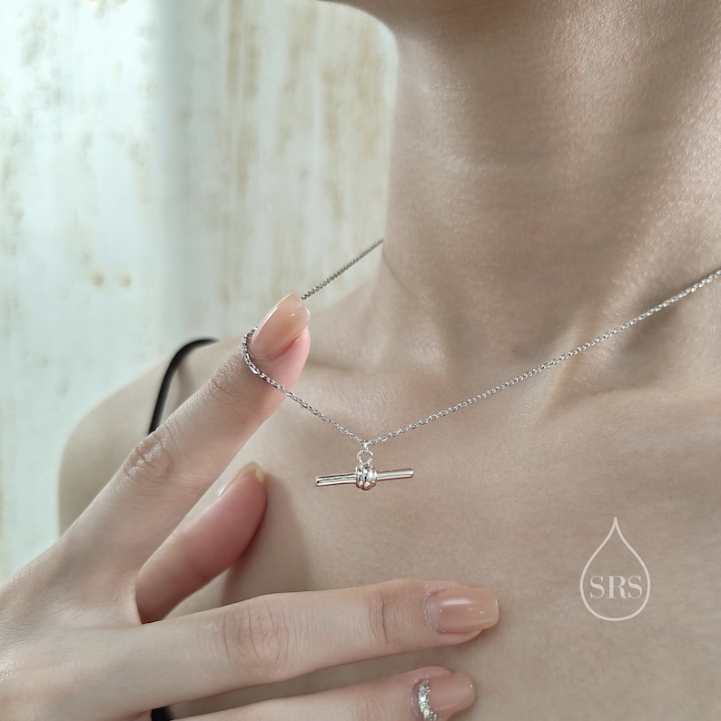 May include: A silver necklace with a toggle clasp pendant. The necklace is on a model's neck and the pendant is in the center of the image.