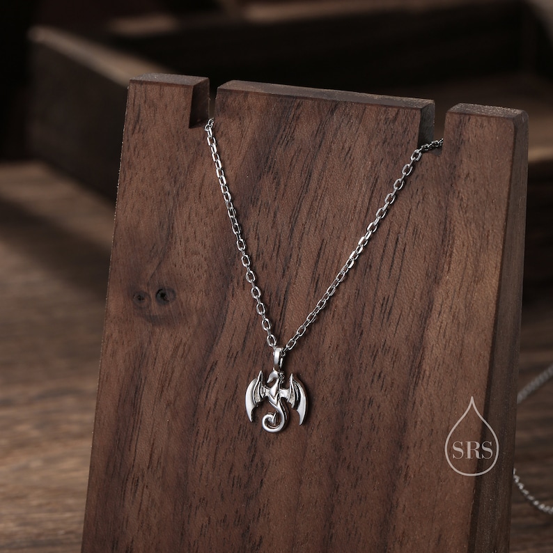 May include: Silver dragon pendant necklace on a silver chain. The pendant is a detailed design of a dragon with wings outstretched. The necklace is displayed on a wooden stand.
