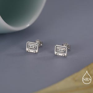 May include: A pair of silver stud earrings in the shape of miniature cassette tapes. The earrings feature detailed rectangular and circular elements, set against a grey background. The design has a retro, vintage aesthetic.
