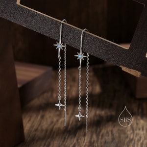 Puede incluir: Un par de pendientes de plata con un diseño de estrella y una piedra de ópalo azul. Los pendientes tienen una cadena larga con una estrella más pequeña al final.