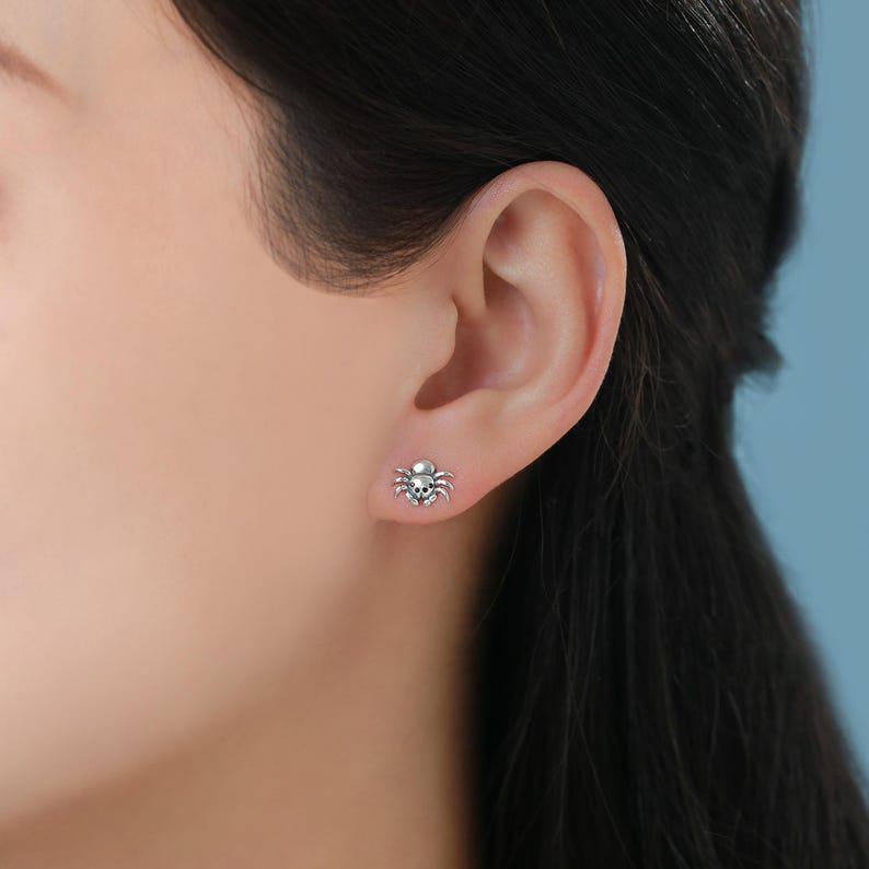 May include: A close-up of a person's ear with a small silver spider stud earring. The spider has a skull-shaped head.