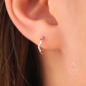 May include: A close-up of a person's ear with a silver wavy hoop earring. The earring is a simple design with a slightly curved shape.