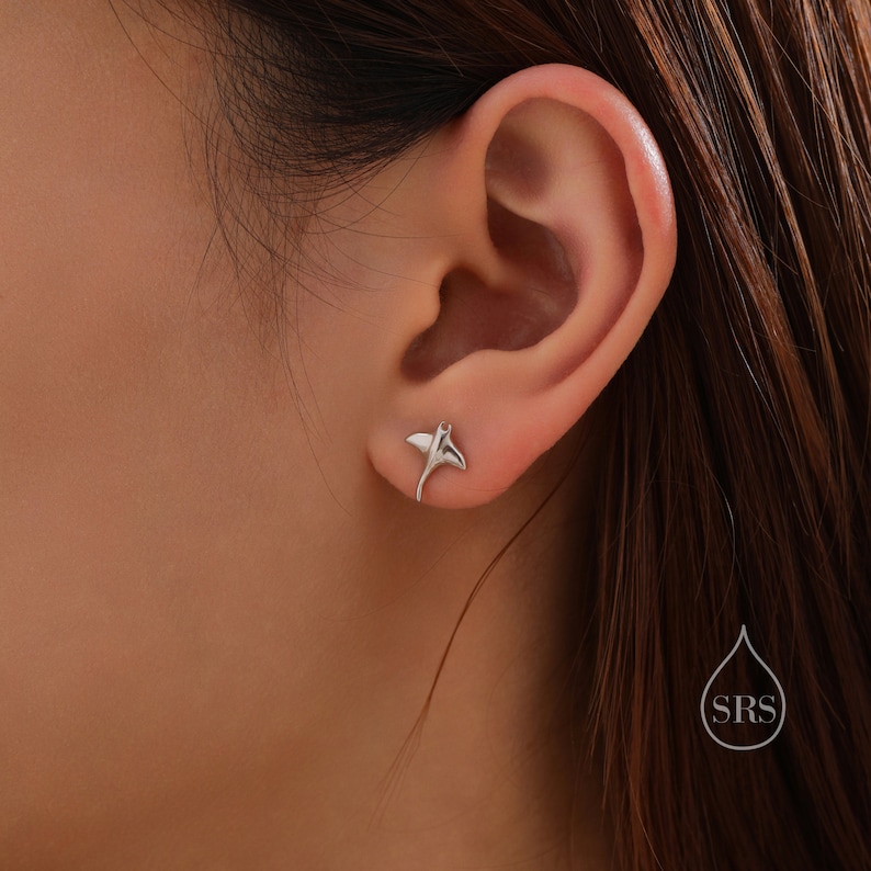 May include: A pair of silver stud earrings featuring a small, detailed stingray design. The earrings are shown in a close-up image of a person's ear.
