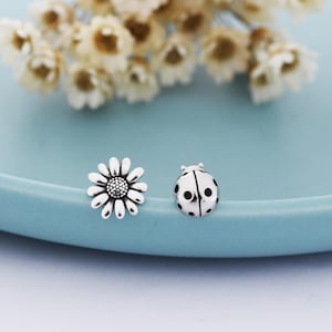 Puede incluir: Pendientes de botón plateados con forma de margarita y mariquita. La margarita tiene pétalos detallados y un centro texturizado. La mariquita presenta manchas negras y una forma redondeada. Los pendientes se muestran sobre una superficie azul claro.