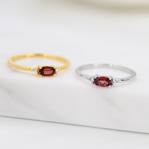 May include: Two rings featuring oval red gemstones and small white stones. One ring is gold-coloured, the other silver. Both are set on a white surface. The rings are suitable for everyday wear.
