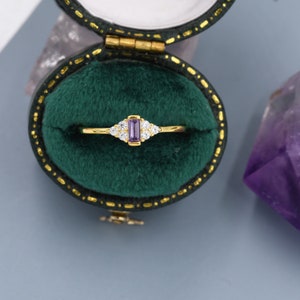 Puede incluir: Un anillo de oro con una piedra rectangular de color p&uacute;rpura rodeada de piedras m&aacute;s peque&ntilde;as de color claro. El anillo est&aacute; en una caja de anillo de terciopelo verde.