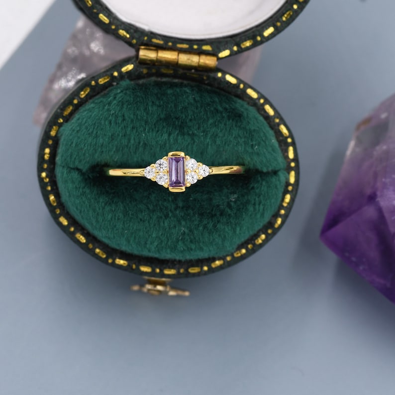 Puede incluir: Un anillo de oro con una piedra rectangular de color p&uacute;rpura rodeada de piedras m&aacute;s peque&ntilde;as de color claro. El anillo est&aacute; en una caja de anillo de terciopelo verde.