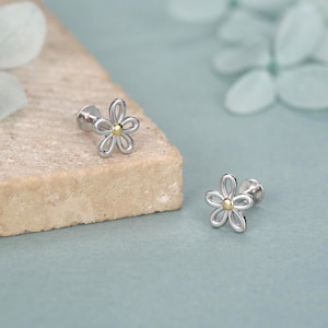 May include: Two silver flower-shaped earrings with a gold center. The earrings are on a light brown surface against a light blue background. The earrings have a screw-back design.